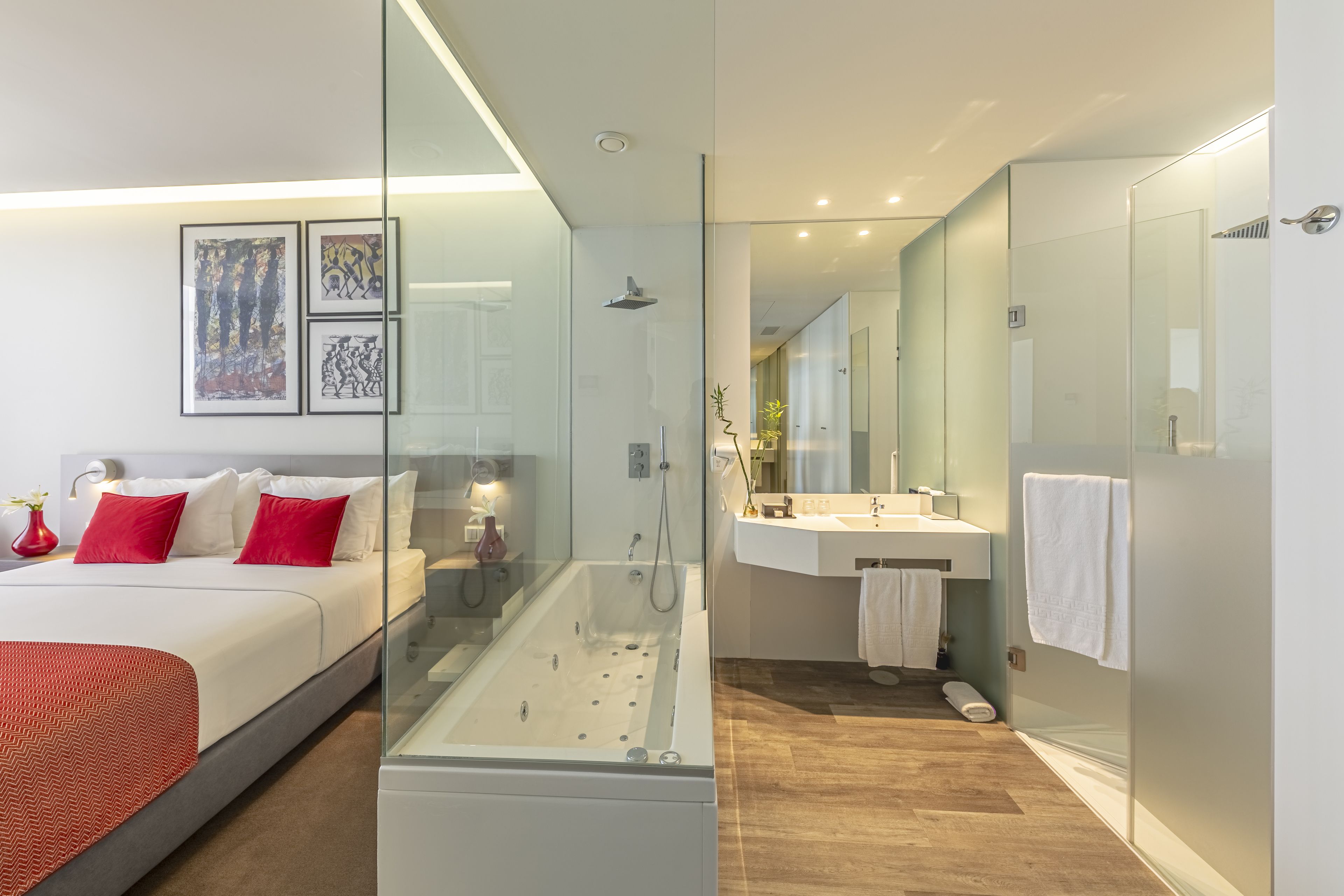 A modern hotel room with a glass-enclosed shower, a large bed with red pillows, a white bedside table, and a mounted artwork above the bed.