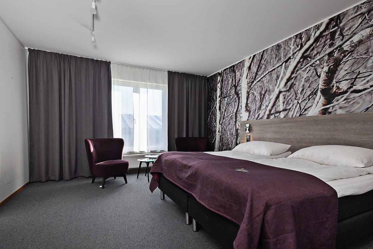 Ett hotellrum med en stor säng, ett skrivbord, två stolar och utsikt över träd genom fönstret.