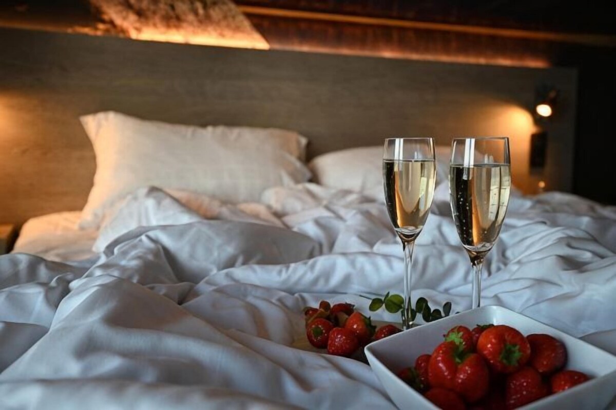Un lit avec du linge de lit blanc, deux coupes de champagne et un bol de fraises.
