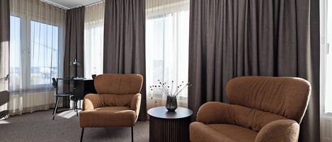 A modern hotel room with two brown armchairs, a round coffee table, and a desk with a chair. Large windows with curtains are visible.