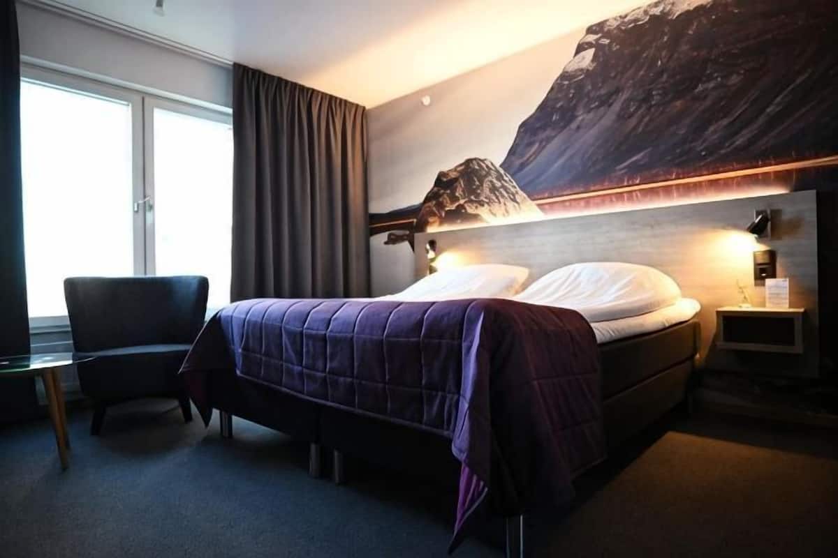 Ett hotellrum med en säng, en stol, ett skrivbord och ett stort fönster.