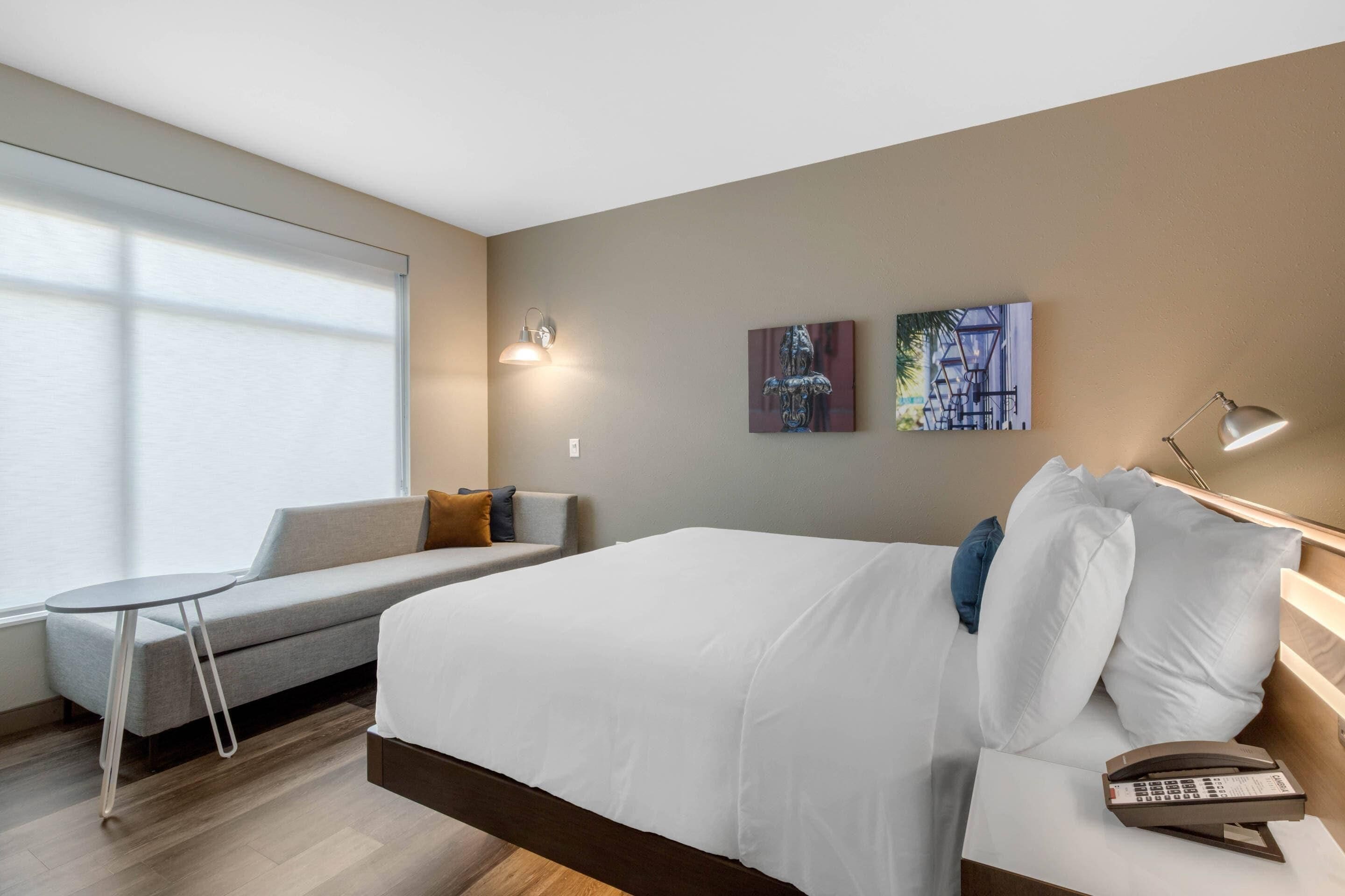 A modern hotel room with a large bed, a small sofa, a bedside table, a lamp, and artwork on the wall.