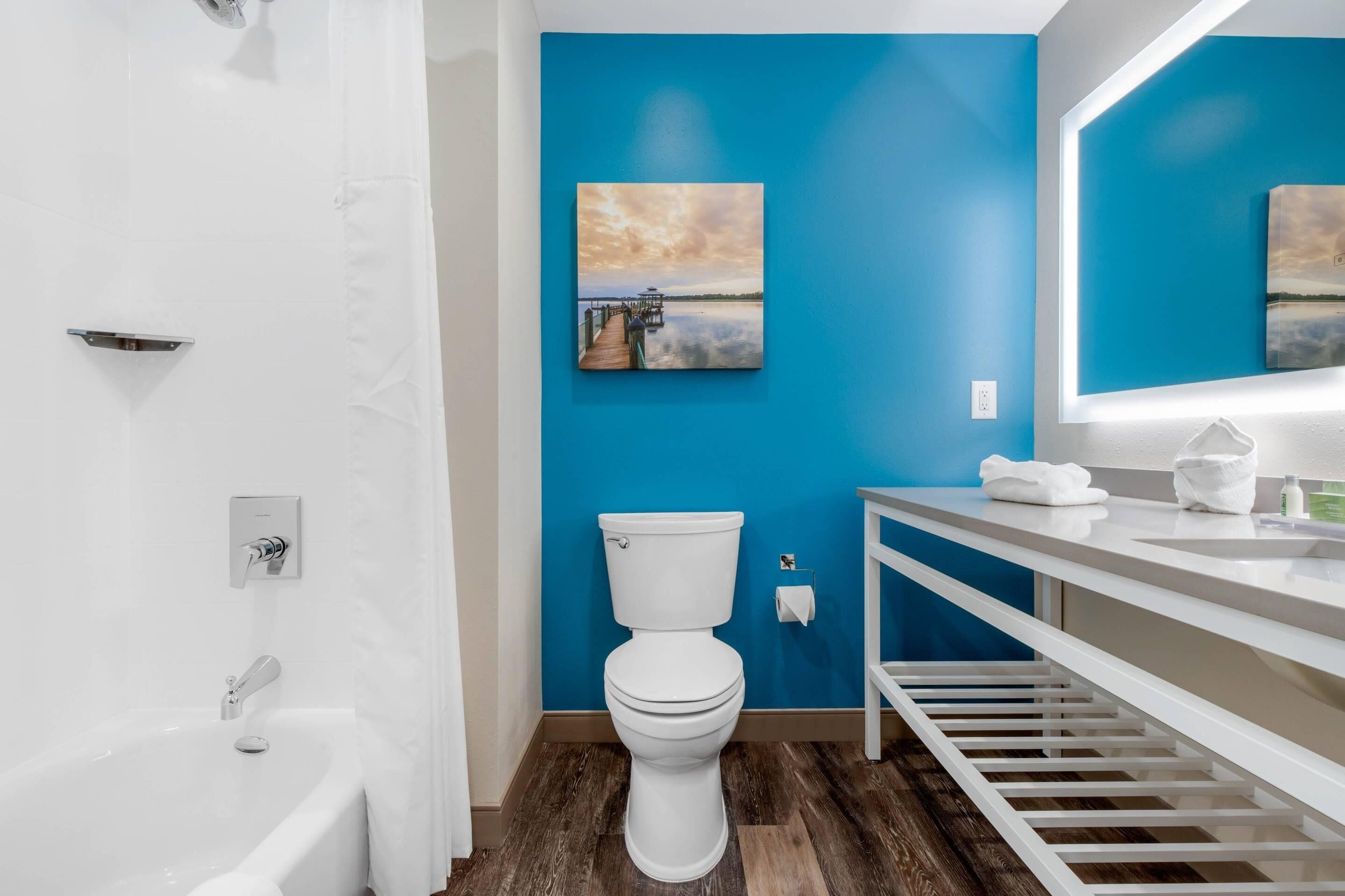 Un baño con inodoro y lavamanos blancos, una pared azul y un cuadro de un atardecer sobre el agua.