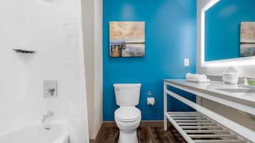 Un baño con inodoro y lavamanos blancos, una pared azul y un cuadro de un atardecer sobre el agua.
