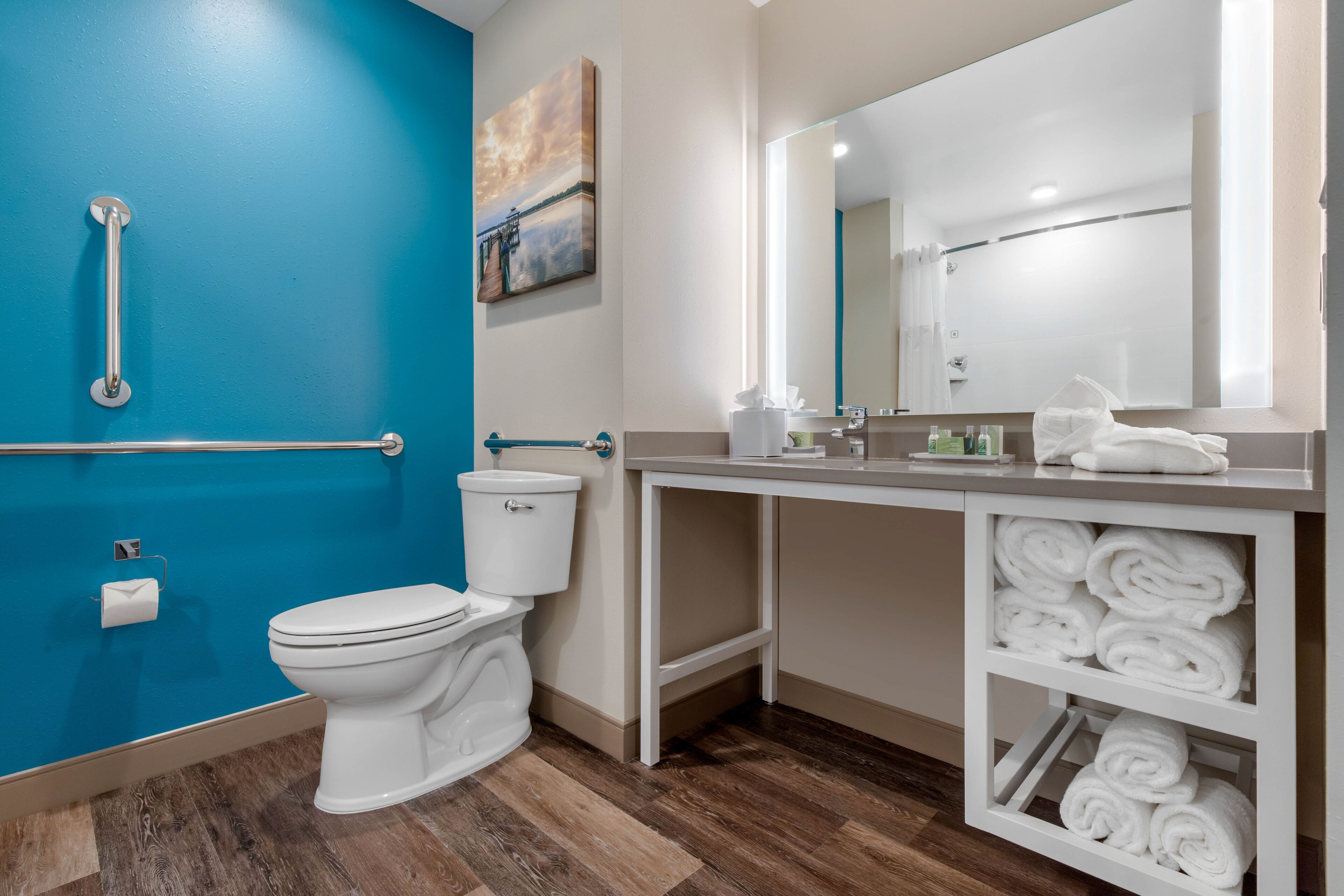 A bathroom with a blue wall, a white toilet, a handrail, a sink with a mirror, and a towel rack.