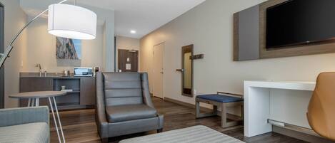 A modern hotel room with a sofa, armchair, ottoman, desk, and a large TV mounted on the wall.
