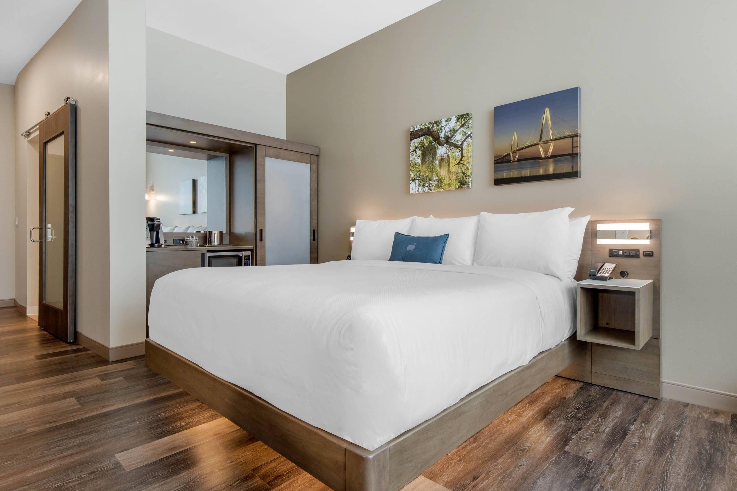 A modern hotel room with a large bed, a built-in wardrobe, a small desk, and a nightstand with a lamp.