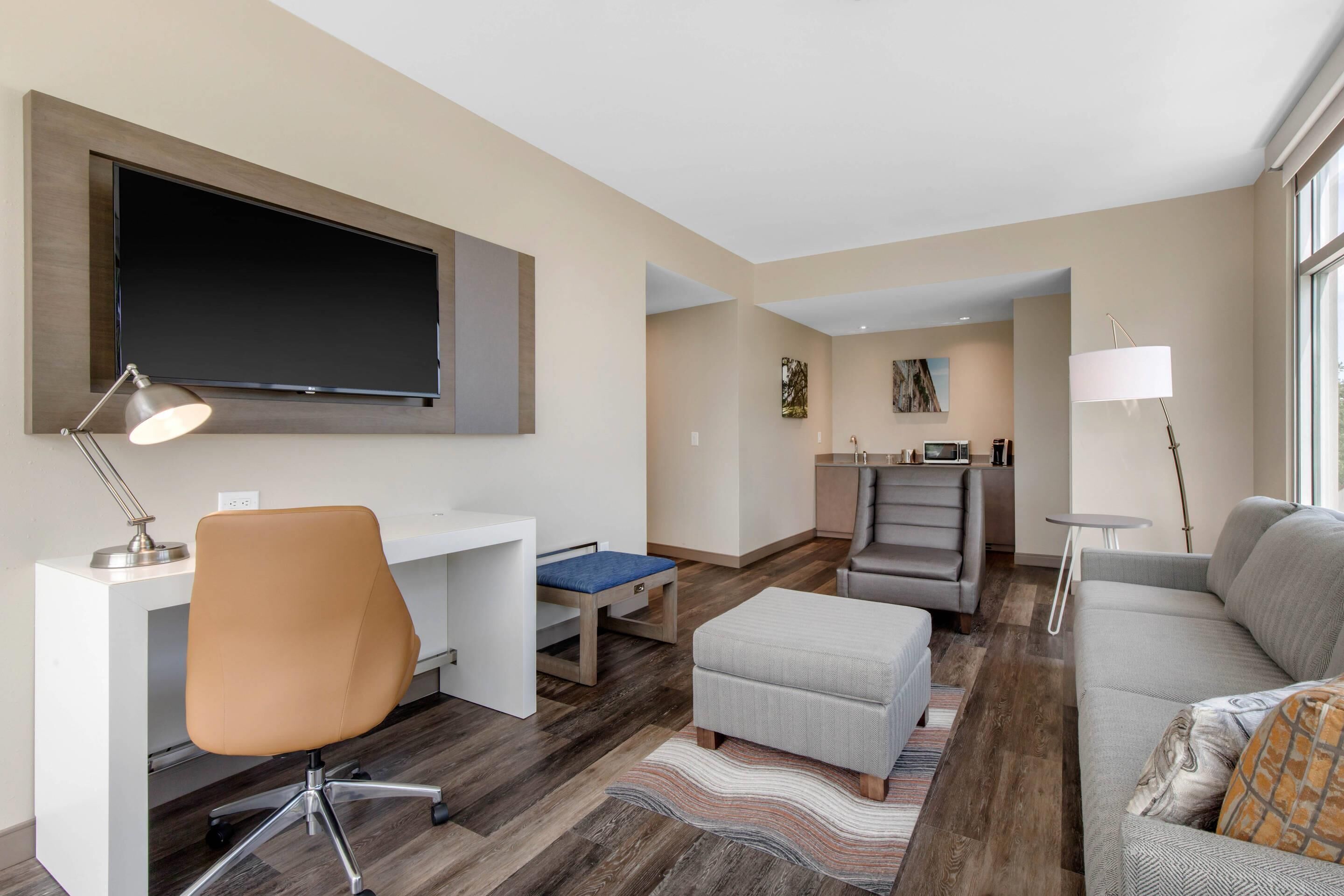 A modern living room with a flat-screen TV, a desk, a comfortable sofa, and a small kitchen area.