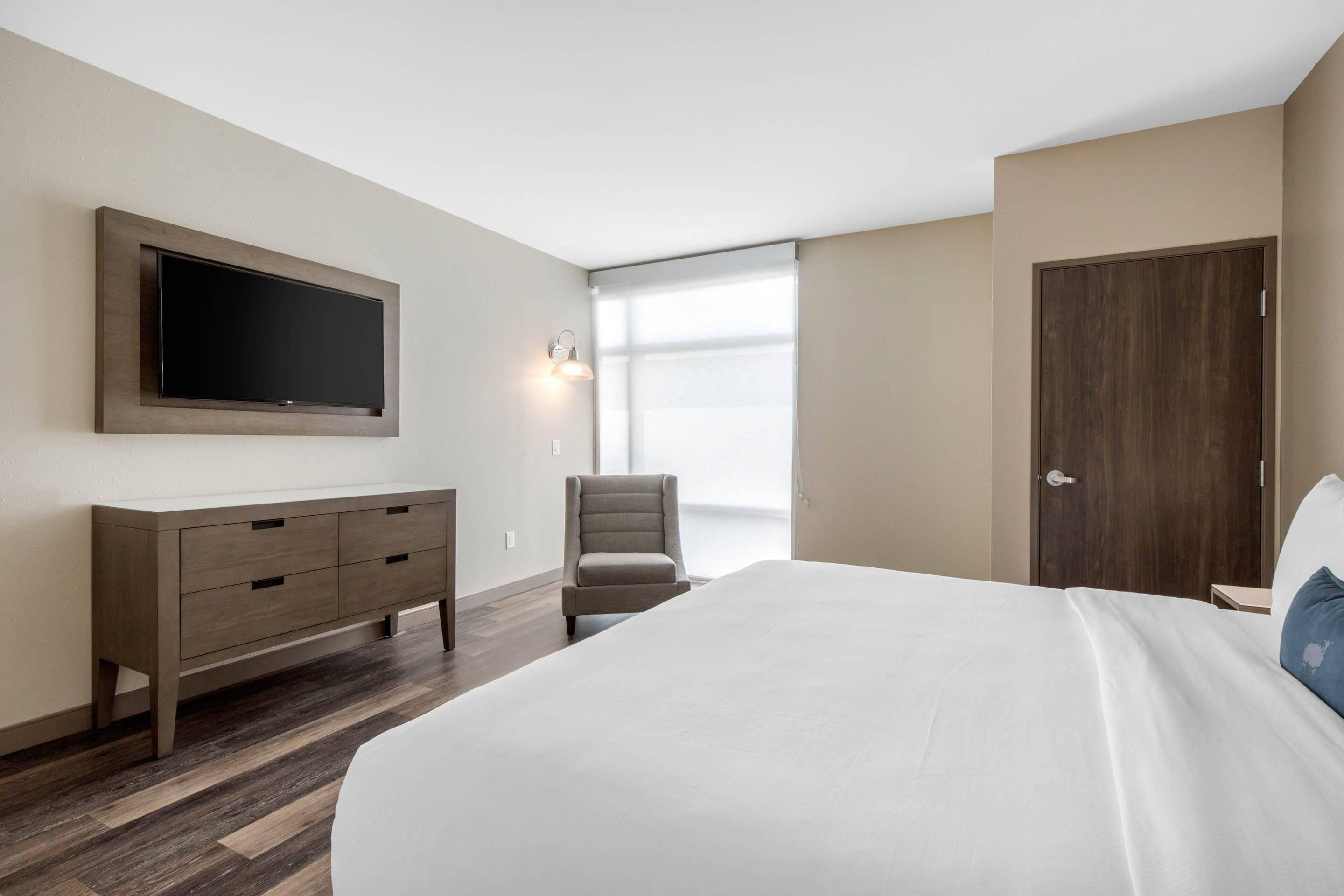 A bedroom with a bed, a TV mounted on the wall, a chair, a dresser, and a door.