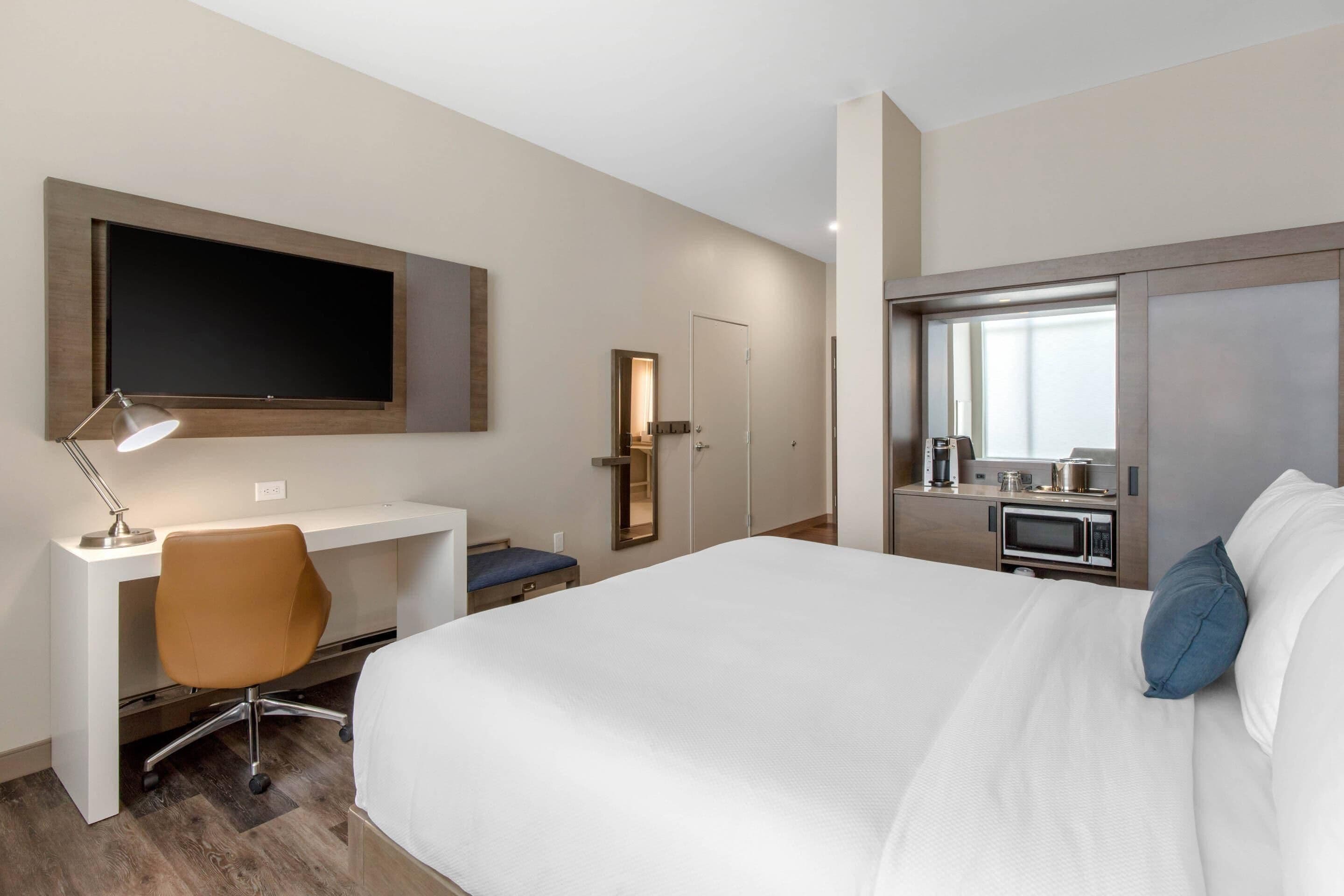 A hotel room with a bed, desk, chair, TV, and kitchenette.