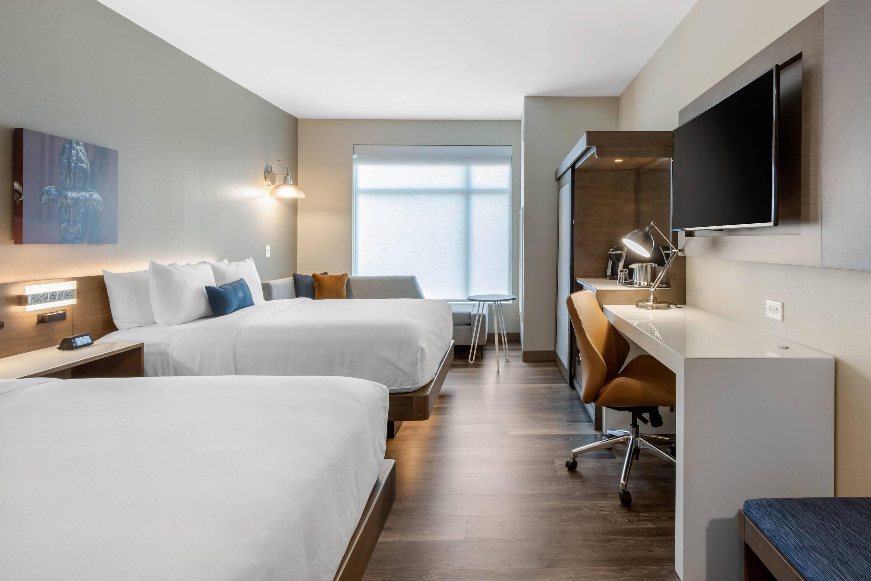 A hotel room with two beds, a desk, a chair, a TV, and a window.