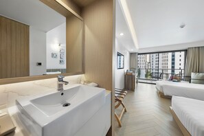 Una habitación de hotel moderna con un lavamanos grande en el baño, un banco de madera, dos camas y un balcón con vistas a la ciudad.