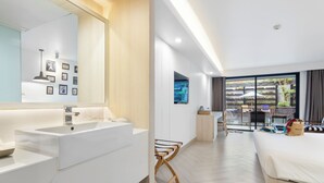 Una habitación de hotel moderna con una cama grande, un espacio de escritorio independiente, un baño con un espejo grande y accesorios blancos, y vistas al exterior a través de ventanas amplias.