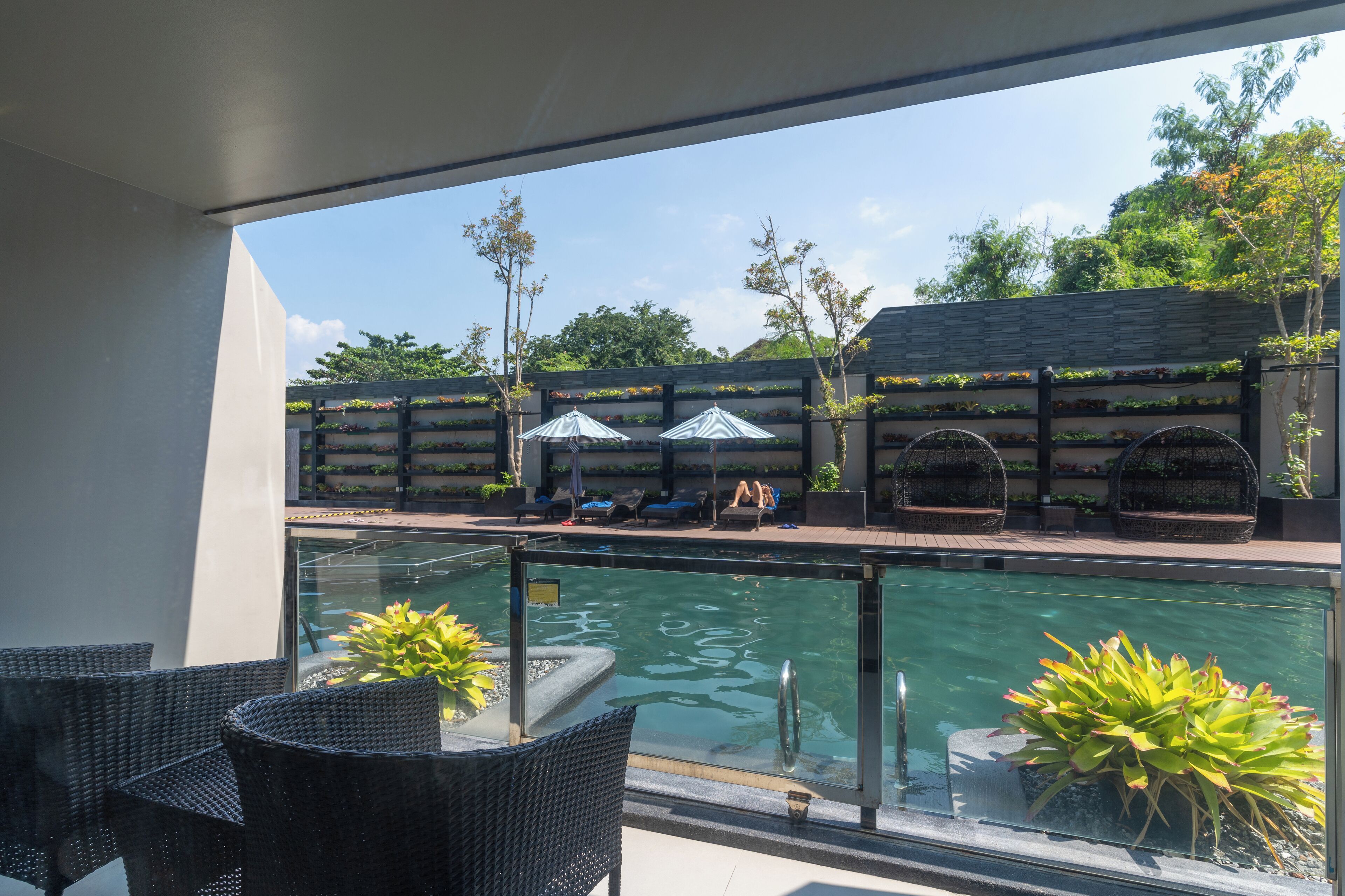 A poolside area with wicker chairs, umbrellas, and a stone wall with planters.