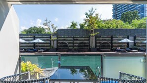 Zona de piscina con sillones reclinables, una mesa de vidrio y vista a un edificio con vegetación y cielo despejado.