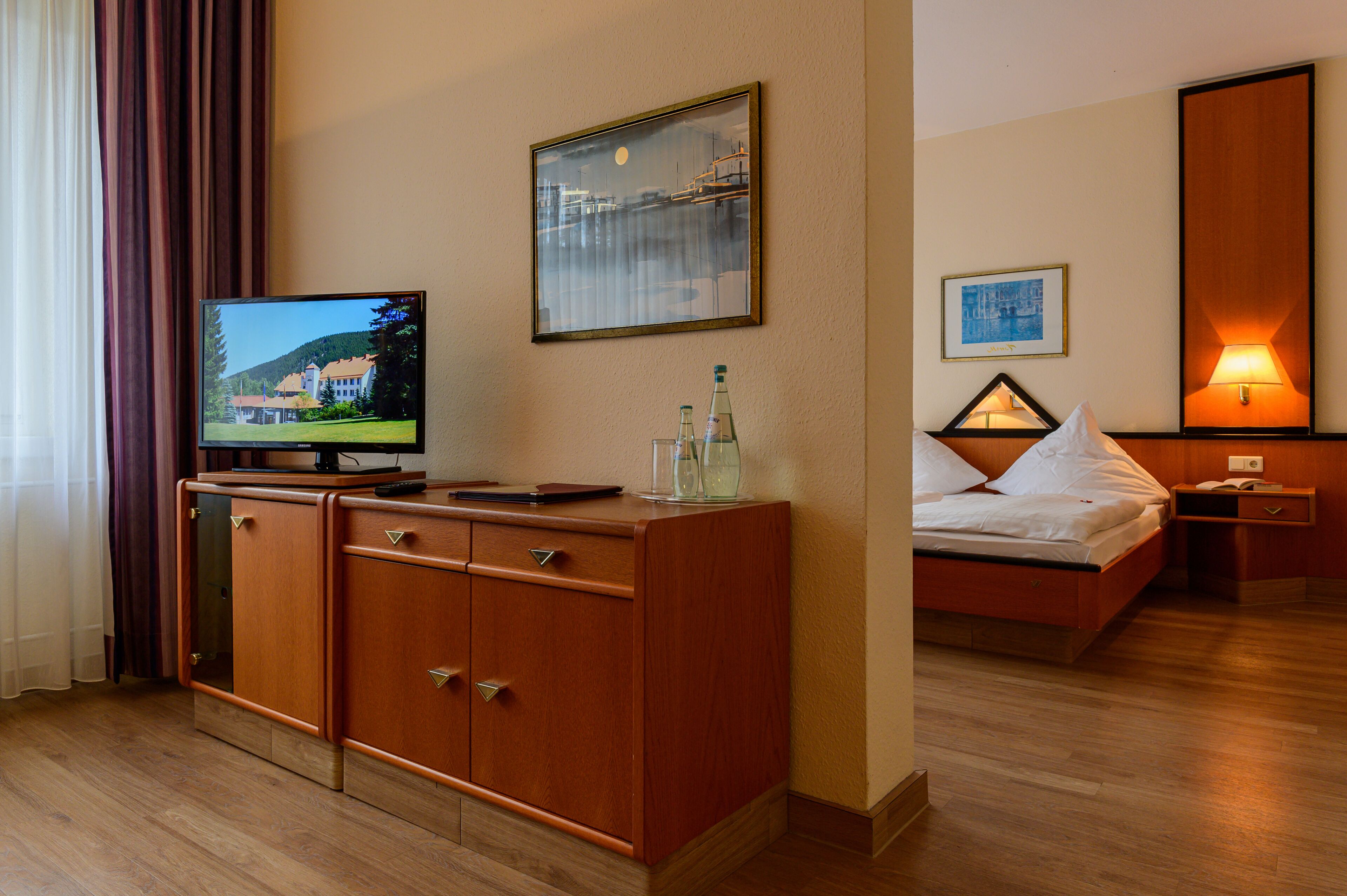 Ein Hotelzimmer mit einem Holzkasten, einem Fernseher, einem Bett mit weißen Leinen und einem Nachttisch, sowie einer Wandlampe.