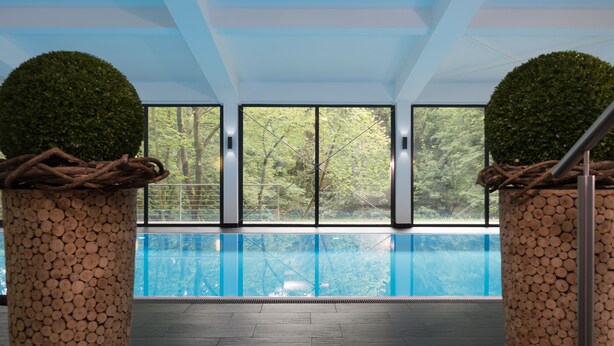 An indoor swimming pool with a view of trees through large windows. - Waldhotel Berghof (Luisenthal)