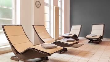 Relax in a serene spa room with four reclining chairs, each with a towel and a small tray, surrounded by large windows and a wall-mounted clock.