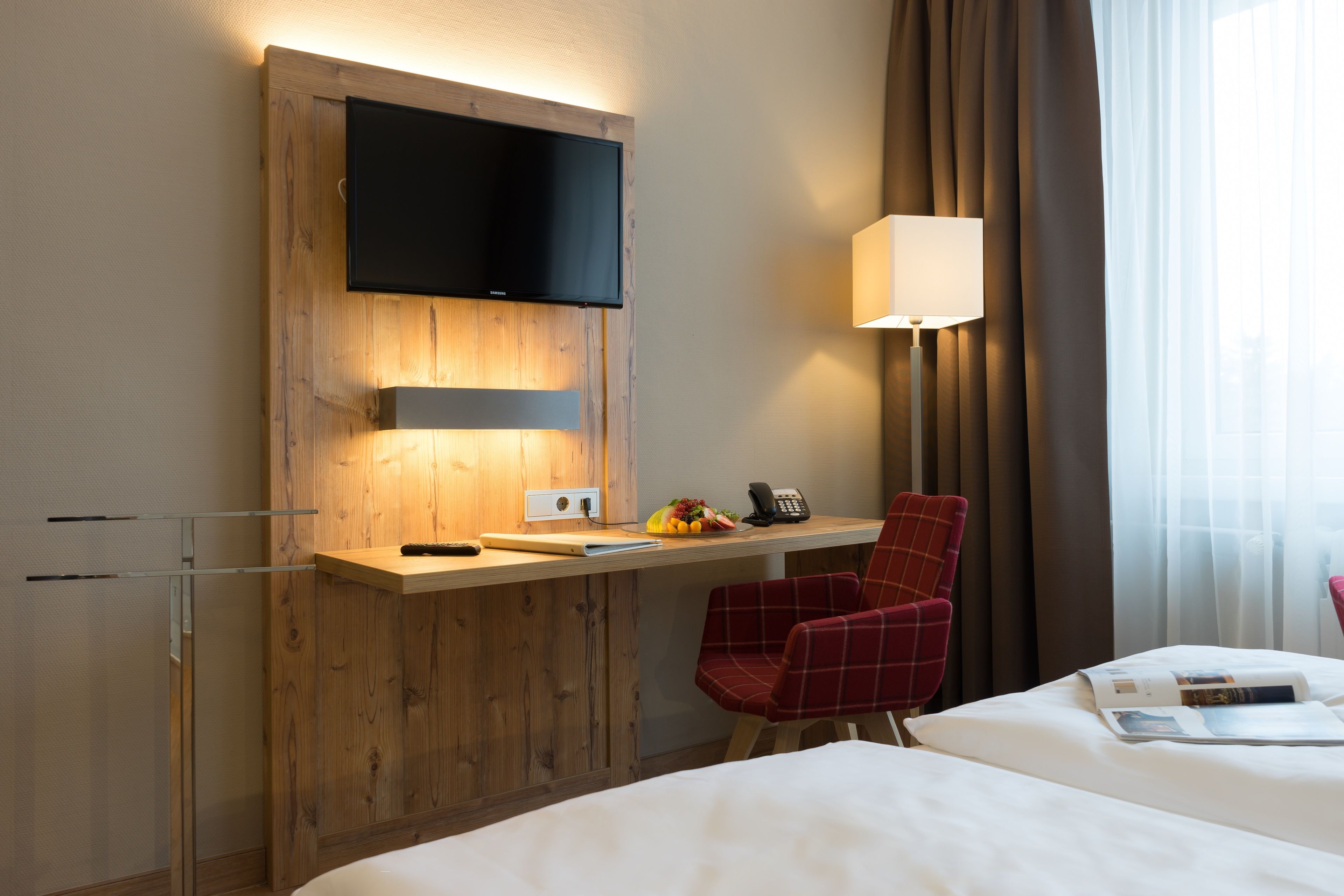 Een hotelkamer met een houten bureau, een flatscreen televisie, een lamp, een rode stoel en een bed met een witte deken.