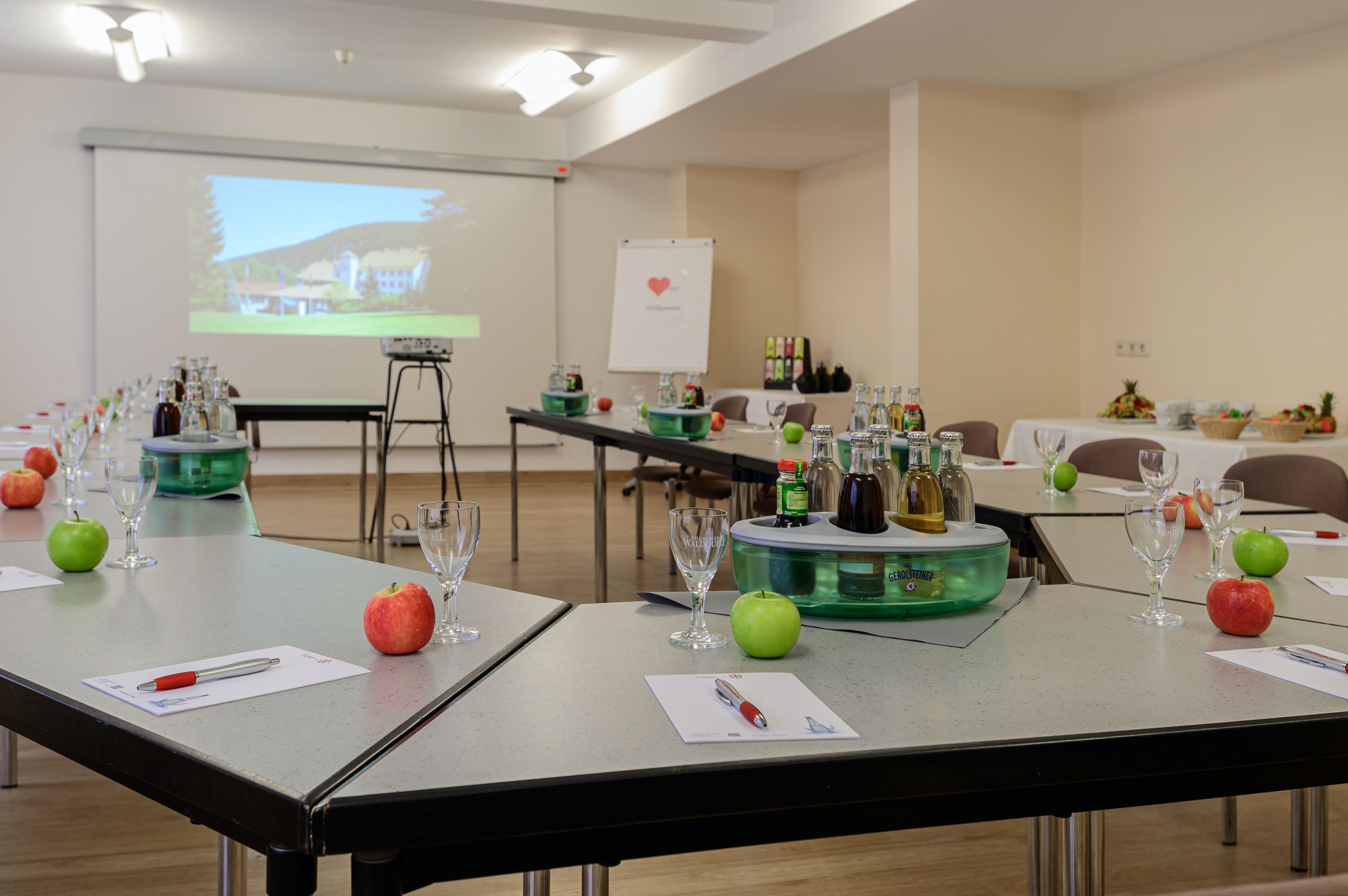 Ein Konferenzraum mit für eine Besprechung gedeckten Tischen, ausgestattet mit Wasserflaschen, Gläsern und Äpfeln.