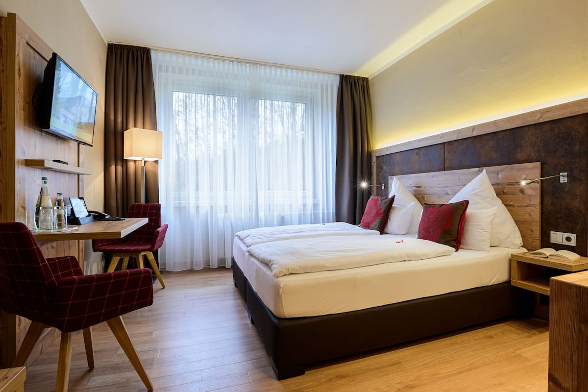A hotel room with a bed, a desk with a chair, a TV, and a window with curtains.