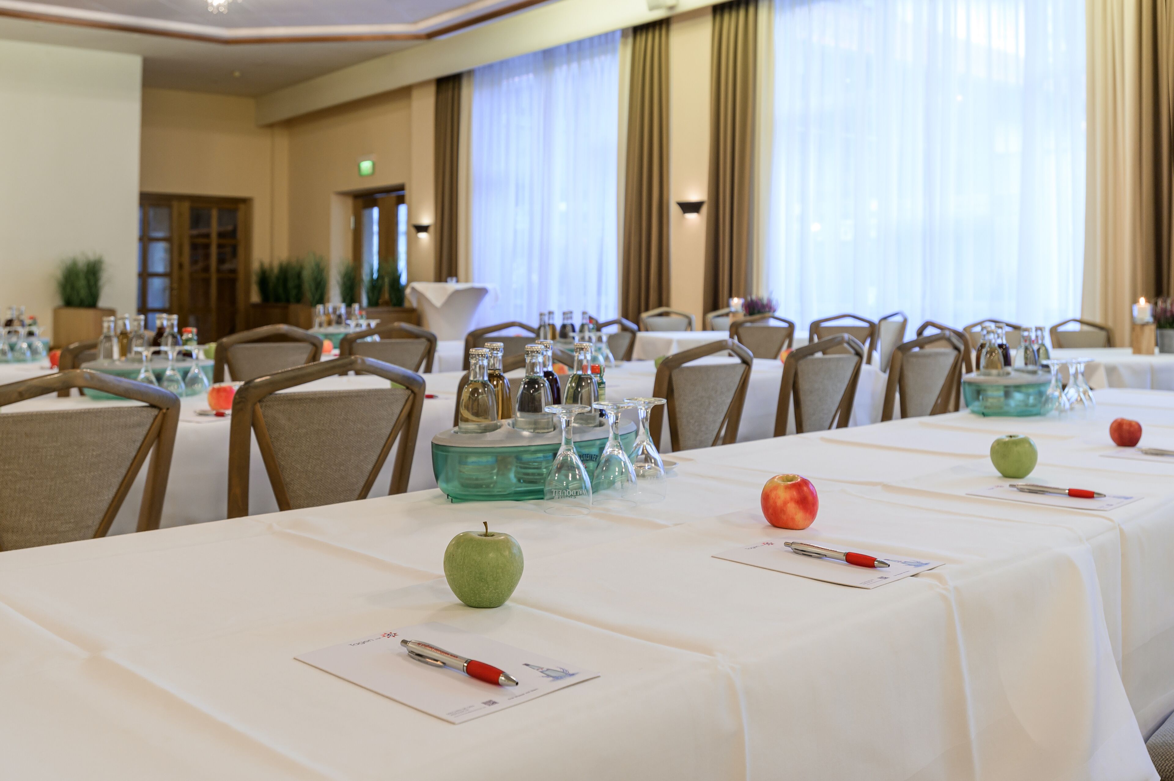 Un salón de conferencias con mesas dispuestas para una reunión, equipado con tarjetas de identificación, bolígrafos y manzanas.
