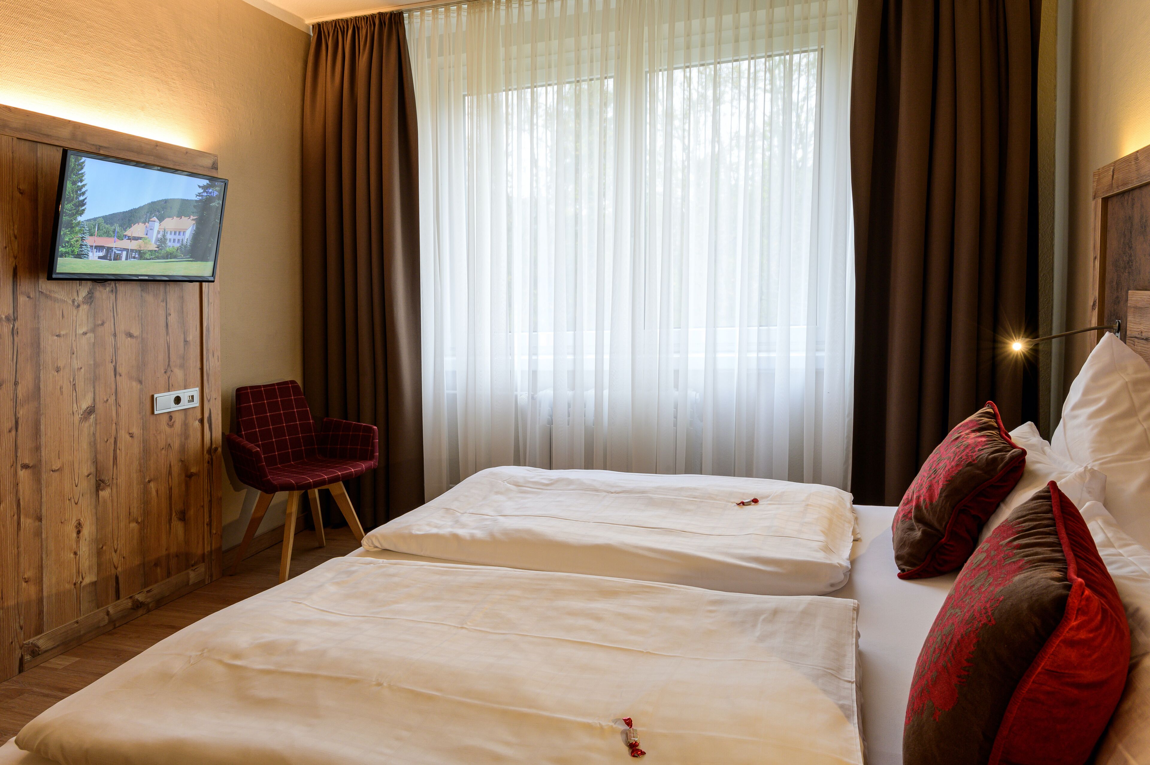 A hotel room with two beds, a TV mounted on a wooden wall, a red chair, and a large window with curtains.