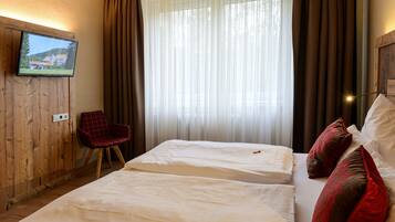 A hotel room with two beds, a TV mounted on a wooden wall, a red chair, and a large window with curtains.