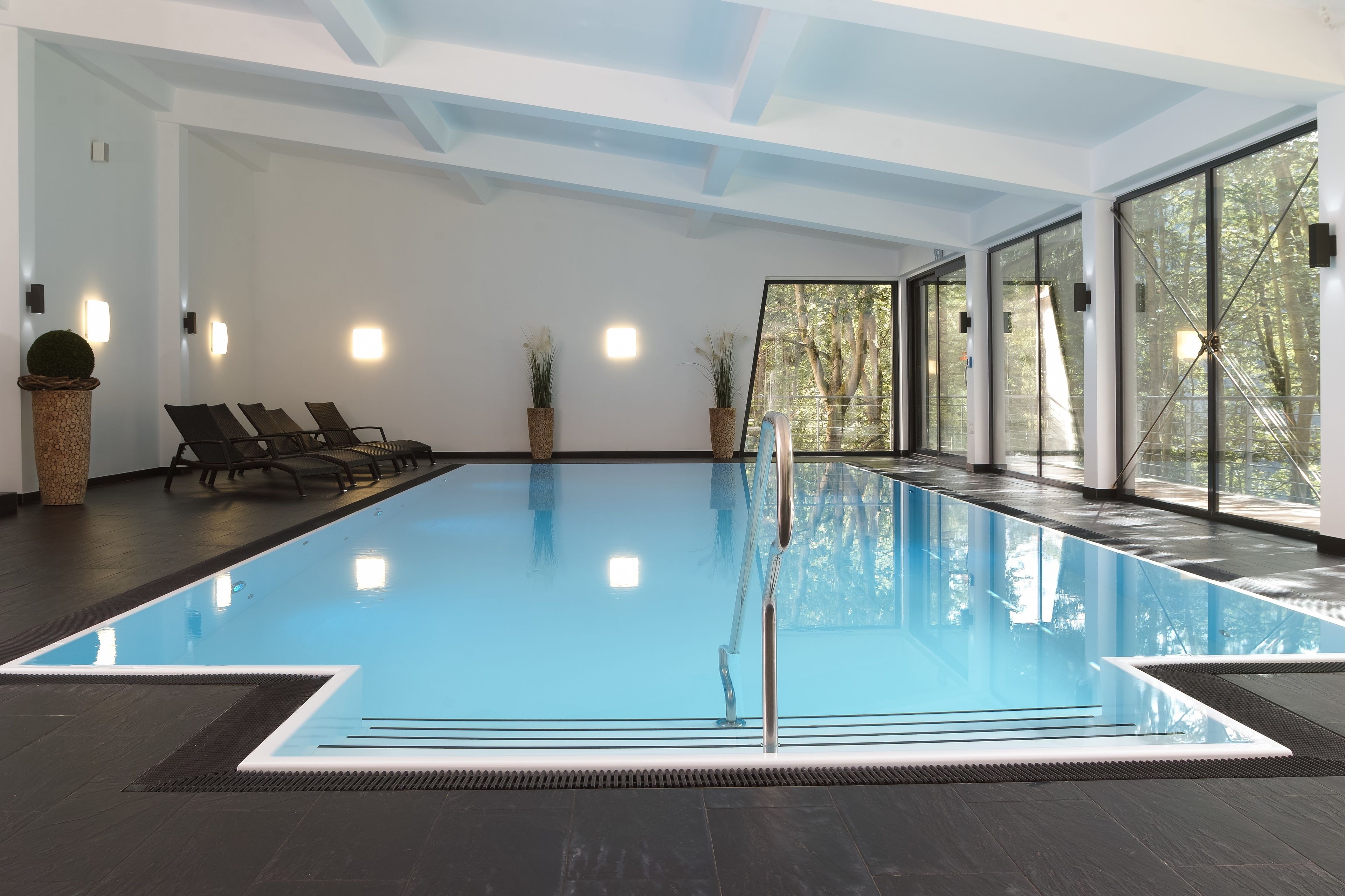 Una amplia piscina interior con paredes de vidrio transparente, suelo oscuro y algunas sillas de descanso.