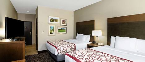 A hotel room with two beds, a desk, a TV, and framed pictures on the wall.
