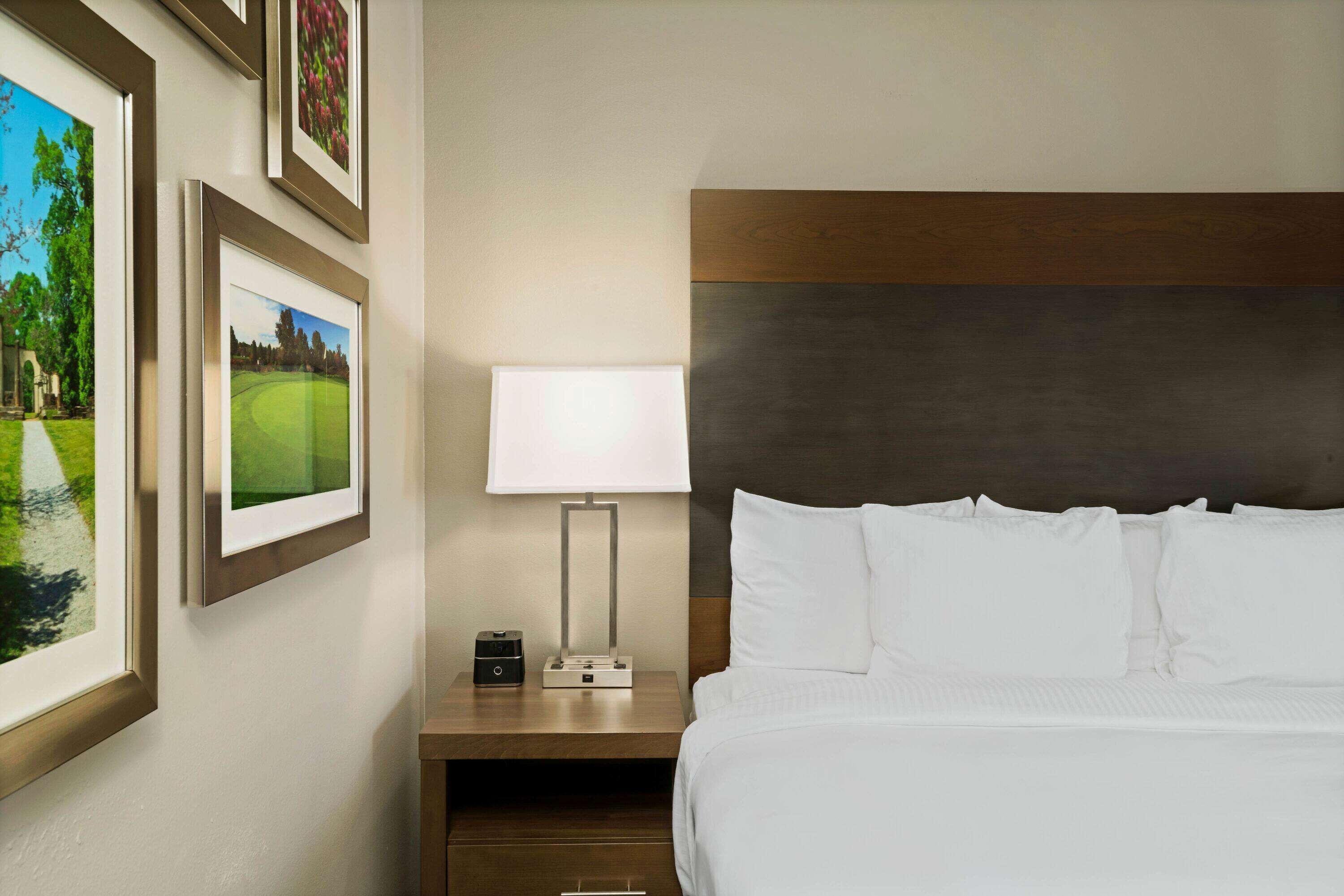 A hotel room with a neatly made bed, a bedside table with a lamp, and framed pictures on the wall.