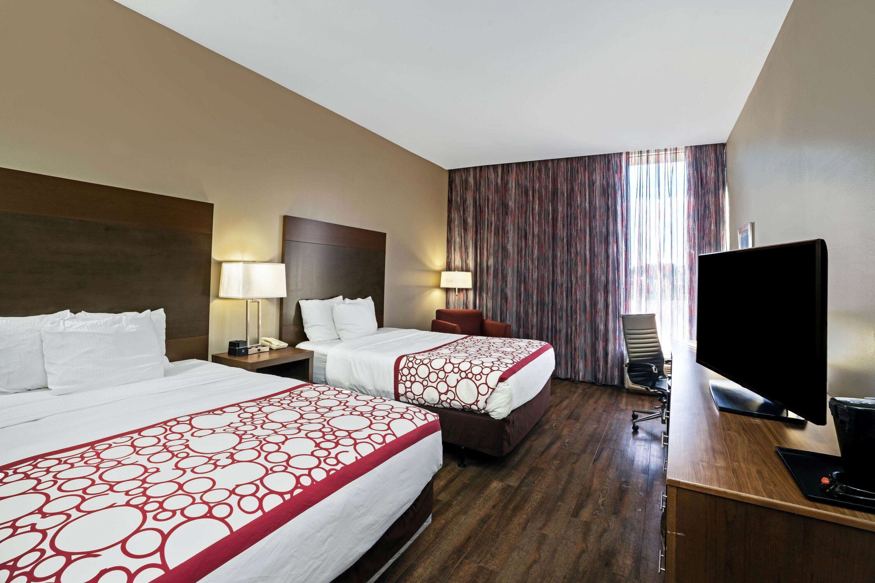 Ein Hotelzimmer mit zwei Betten, einem Schreibtisch mit Computer, einem Fernseher und einem großen Fenster mit Vorhängen.