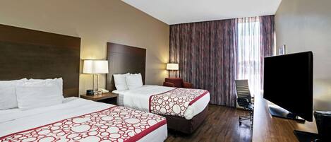 A hotel room with two beds, a desk with a computer, a TV, and a large window with curtains.