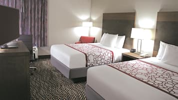 Quarto de hotel com duas camas, uma escrivaninha, uma cadeira, uma televisão e um tapete estampado.