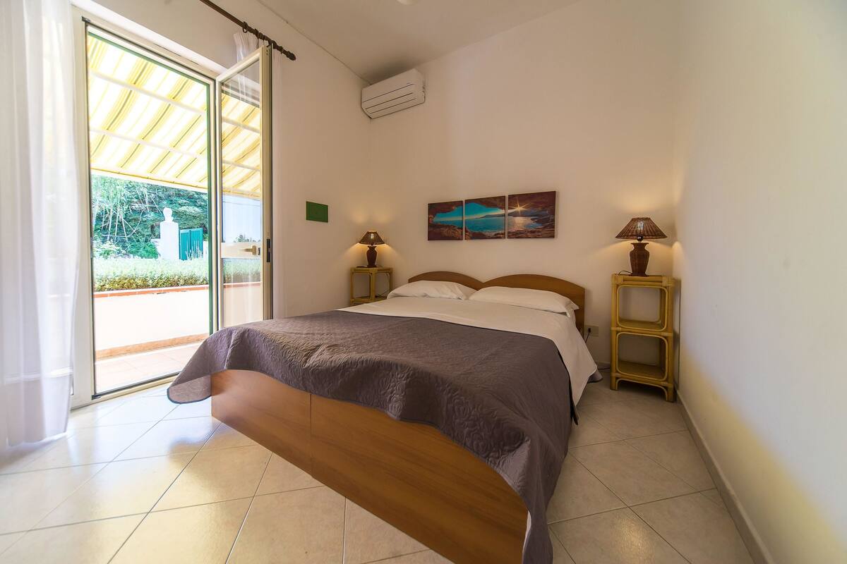 Une chambre à coucher avec un lit, deux tables de chevet avec des lampes, un tableau au mur et une porte-fenêtre coulissante donnant sur l’extérieur.