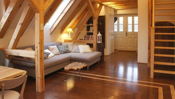 A cozy attic room with a wooden ceiling, a gray sofa, a small table, and a staircase. - Zum Alten Backhaus (Burg (Spreewald))