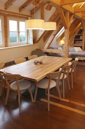 A wooden dining table with chairs, a hanging light fixture, and a view of a window and a sofa in a room with wooden beams. - Zum Alten Backhaus (Burg (Spreewald))
