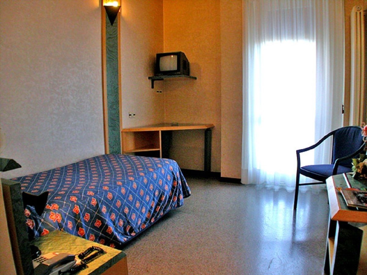 Une chambre d’hôtel avec un lit, un bureau, une chaise, un téléviseur posé sur une étagère et une fenêtre avec des rideaux.
