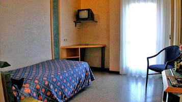 Une chambre d’hôtel avec un lit, un bureau, une chaise, un téléviseur posé sur une étagère et une fenêtre avec des rideaux.