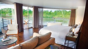 A spacious bedroom with a large bed, a glass table, a chair, and a view of a river. - Jungle Experiences Zafiro (Nauta)