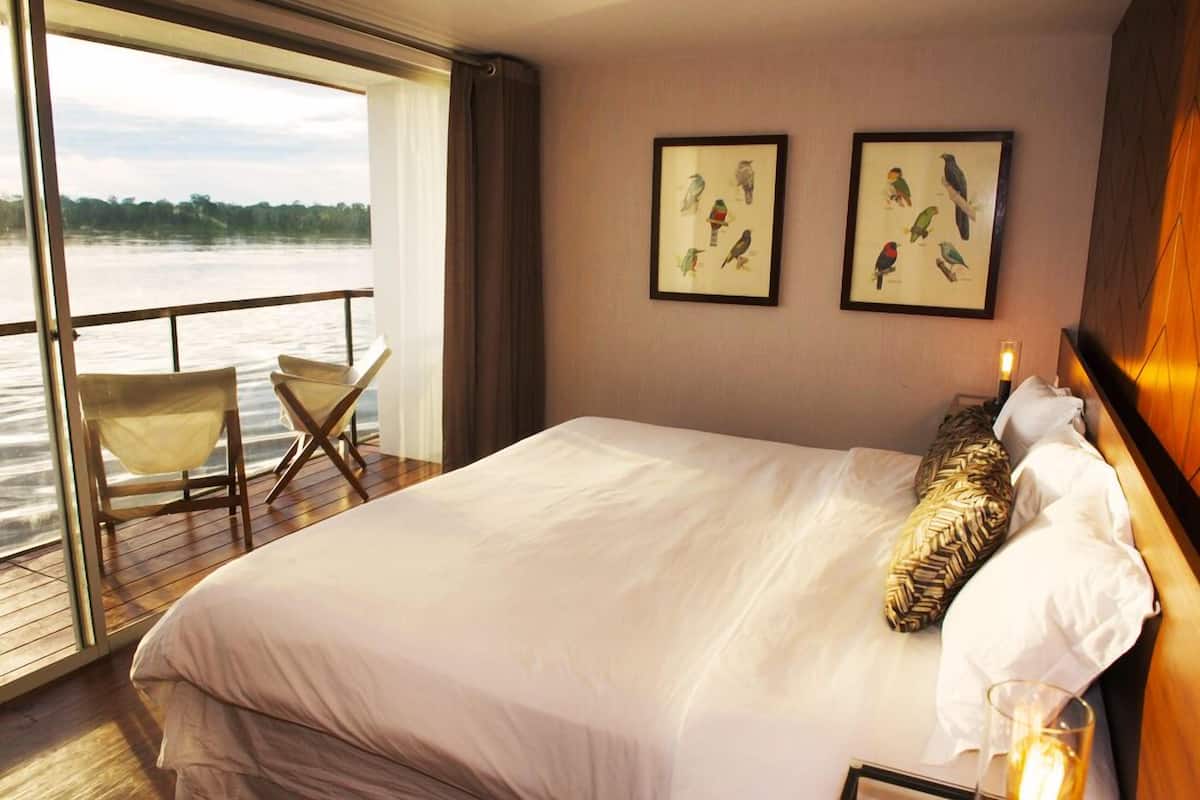 Habitación de hotel con una cama grande, vista a un cuerpo de agua, dos cuadros enmarcados de pájaros en la pared y una silla junto a la ventana.