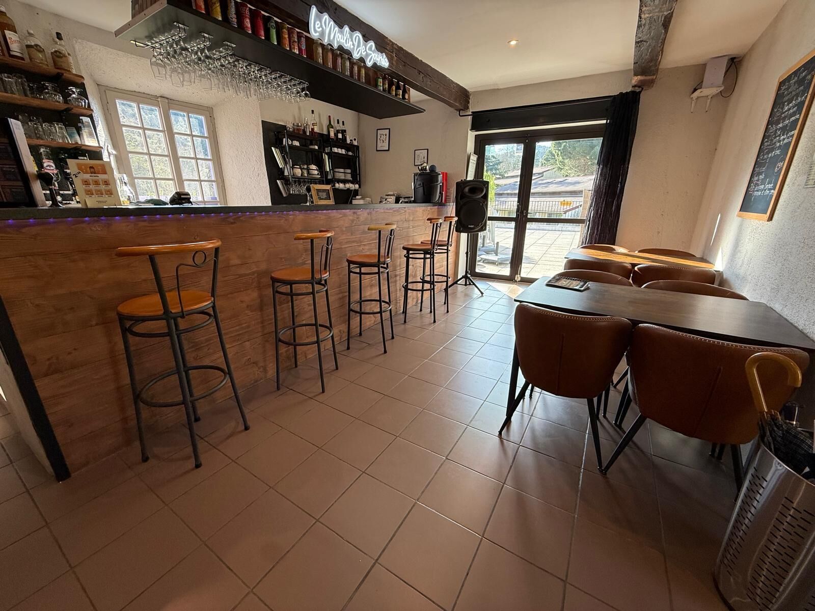 Una zona de bar con taburetes, una barra llena de botellas y vistas por la puerta.
