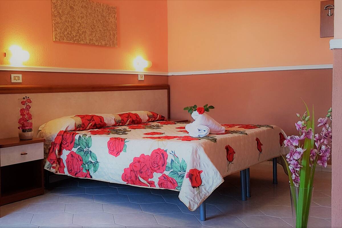 A bedroom with a bed, a nightstand, a wall-mounted light, and a vase with flowers.