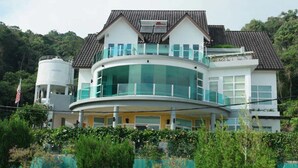 A two-story house with a curved balcony, a water tank, and a garden in the foreground.