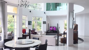 A spacious living area with a high ceiling, large windows, a central seating area with a TV, and a dining table with chairs.