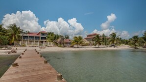 A wooden pier leading to a beach with clear water, palm trees, and a resort building with a red roof. - Villa Mission Bay (Placencia)