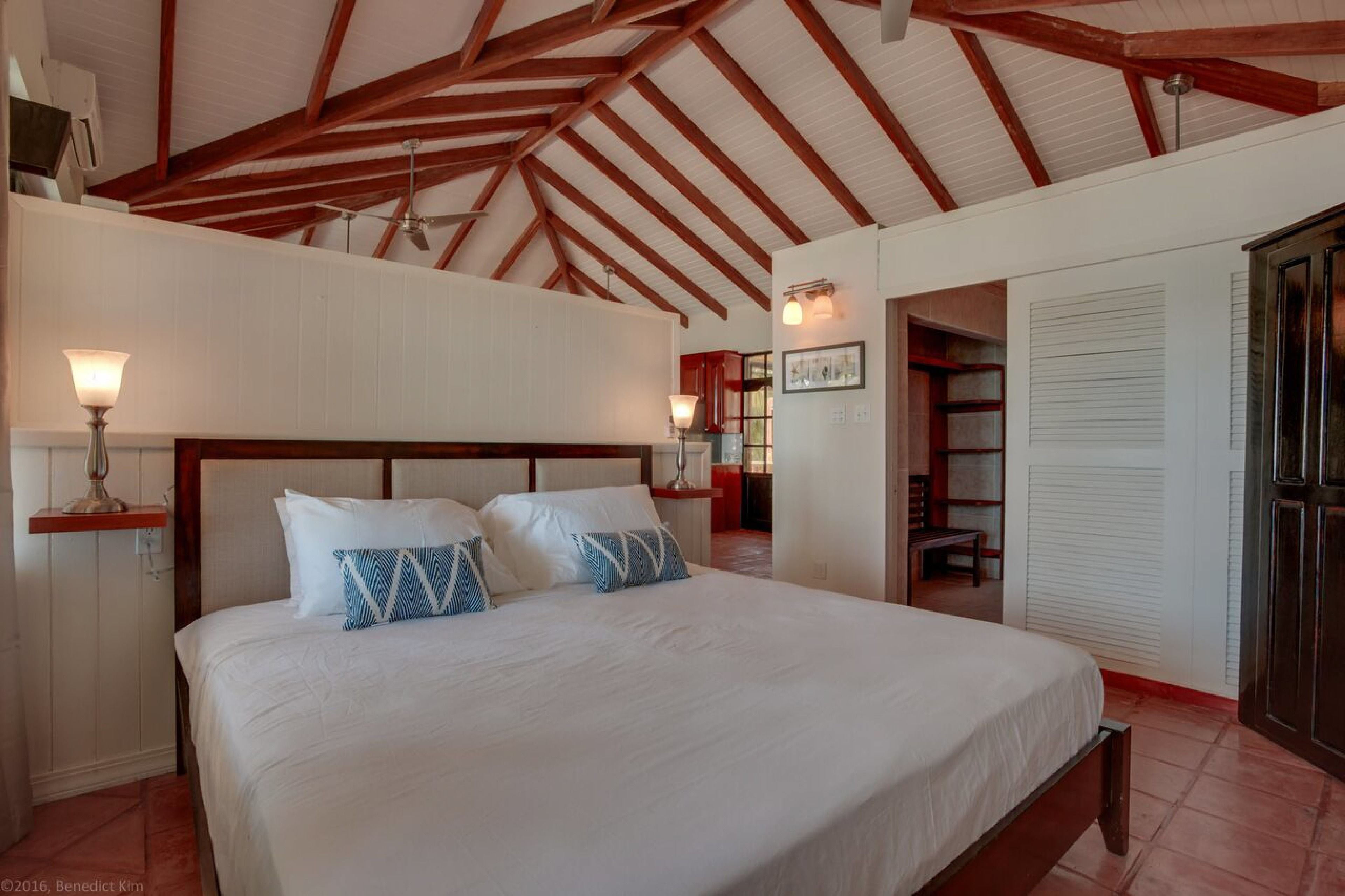 A bedroom with a large bed, wooden beams on the ceiling, a bedside table with a lamp, and a wardrobe.