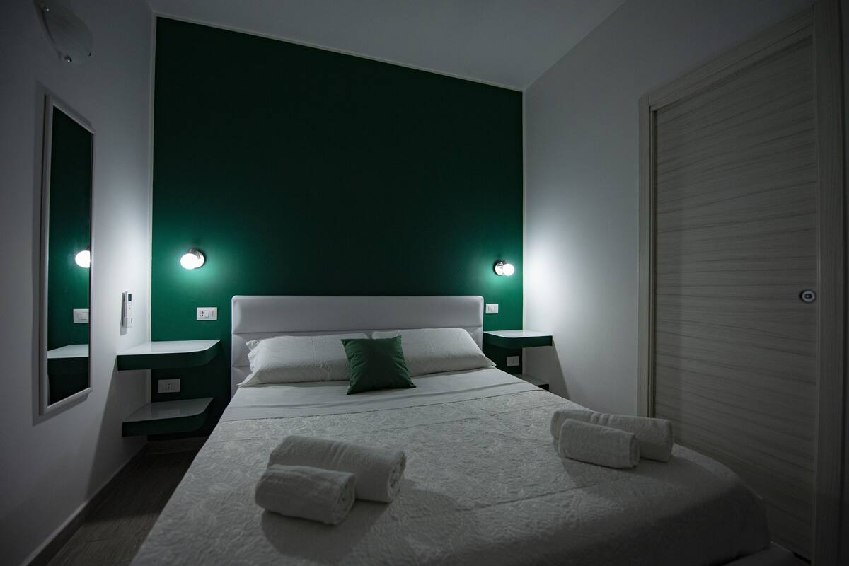 Una cama bien tendida con sábanas blancas y almohadas verdes, dos mesitas de noche con lámparas y una puerta cerrada.