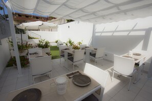 A patio area with white wicker furniture, a table with plates and a mug, and a covered seating area with a view of a building and a garden. - Residenza Magnolia (Tropea)