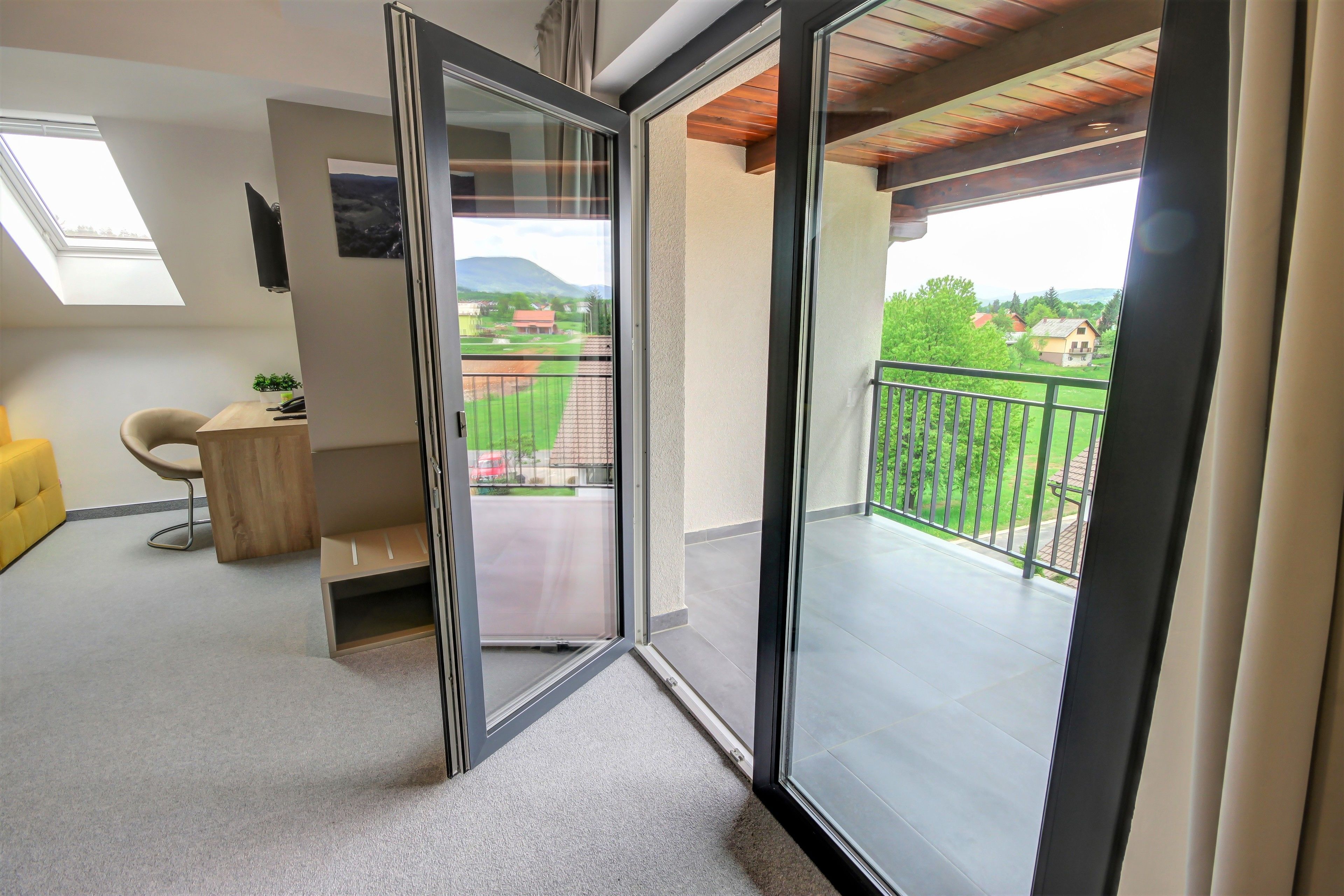 A modern apartment with a sliding glass door leading to a balcony with a view of a green landscape and houses.